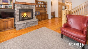 A living room with a stone fireplace, wooden shelves, a red leather chair, and a flat-screen TV. - Inviting Spanish Peaks Townhome|Private Hot Tub| 15 min Drive to Resort (Big Sky)