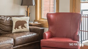 A brown leather sofa with a bear-shaped pillow and a red leather armchair in a room with a wooden floor, a lamp, and a window with a view of a wooden deck. - Inviting Spanish Peaks Townhome|Private Hot Tub| 15 min Drive to Resort (Big Sky)