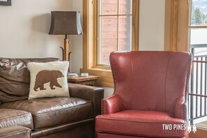 A brown leather sofa with a bear-shaped pillow and a red leather armchair in a room with a wooden floor, a lamp, and a window with a view of a wooden deck. - Inviting Spanish Peaks Townhome|Private Hot Tub| 15 min Drive to Resort (Big Sky)