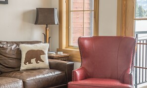 Un sofá de cuero marrón con un cojín con forma de oso y un sillón de cuero rojo en una habitación con piso de madera, una lámpara y una ventana con vista a una terraza de madera.