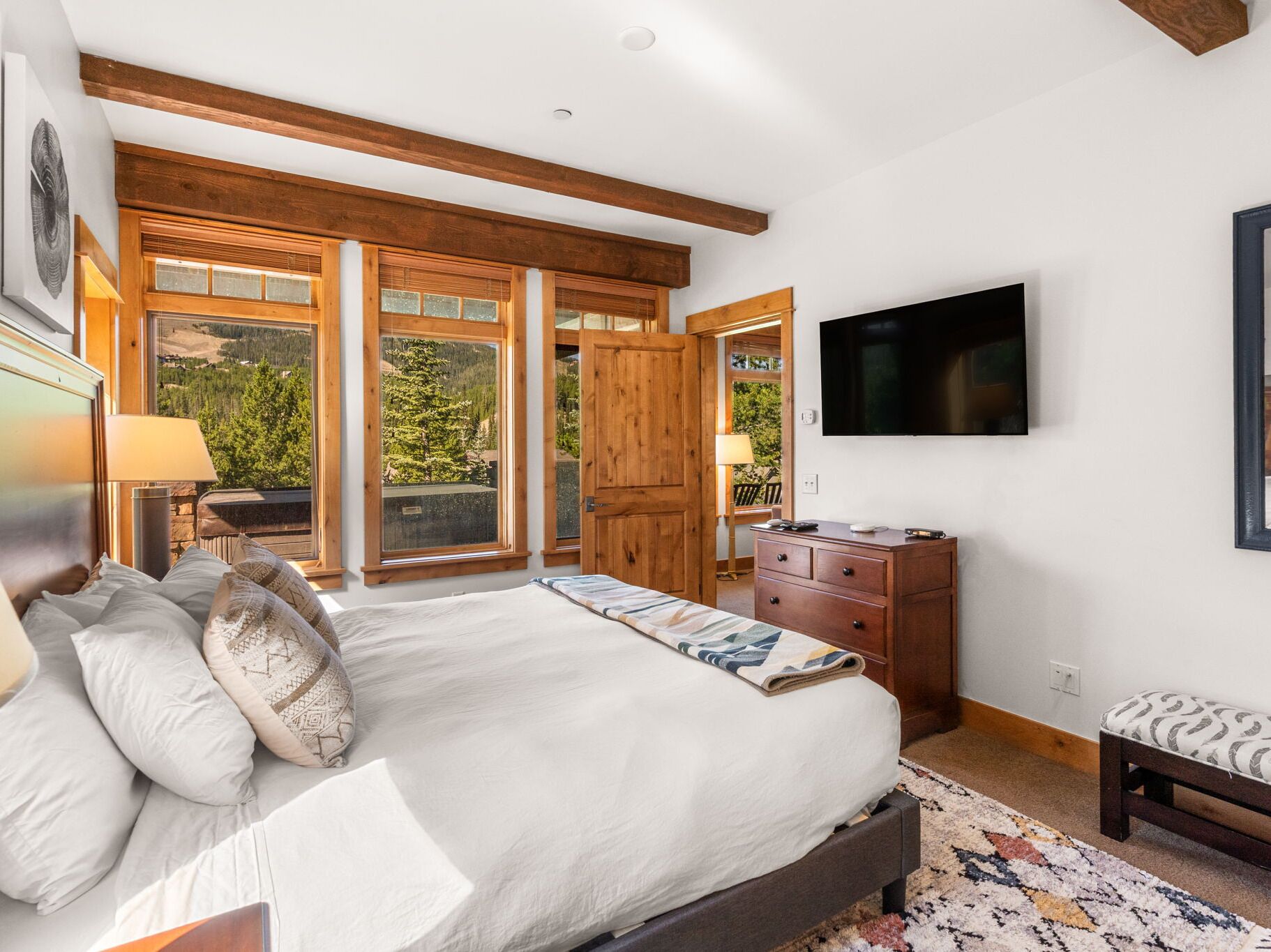 A bedroom with a large bed, wooden beams, a TV, and a view of trees through the windows.