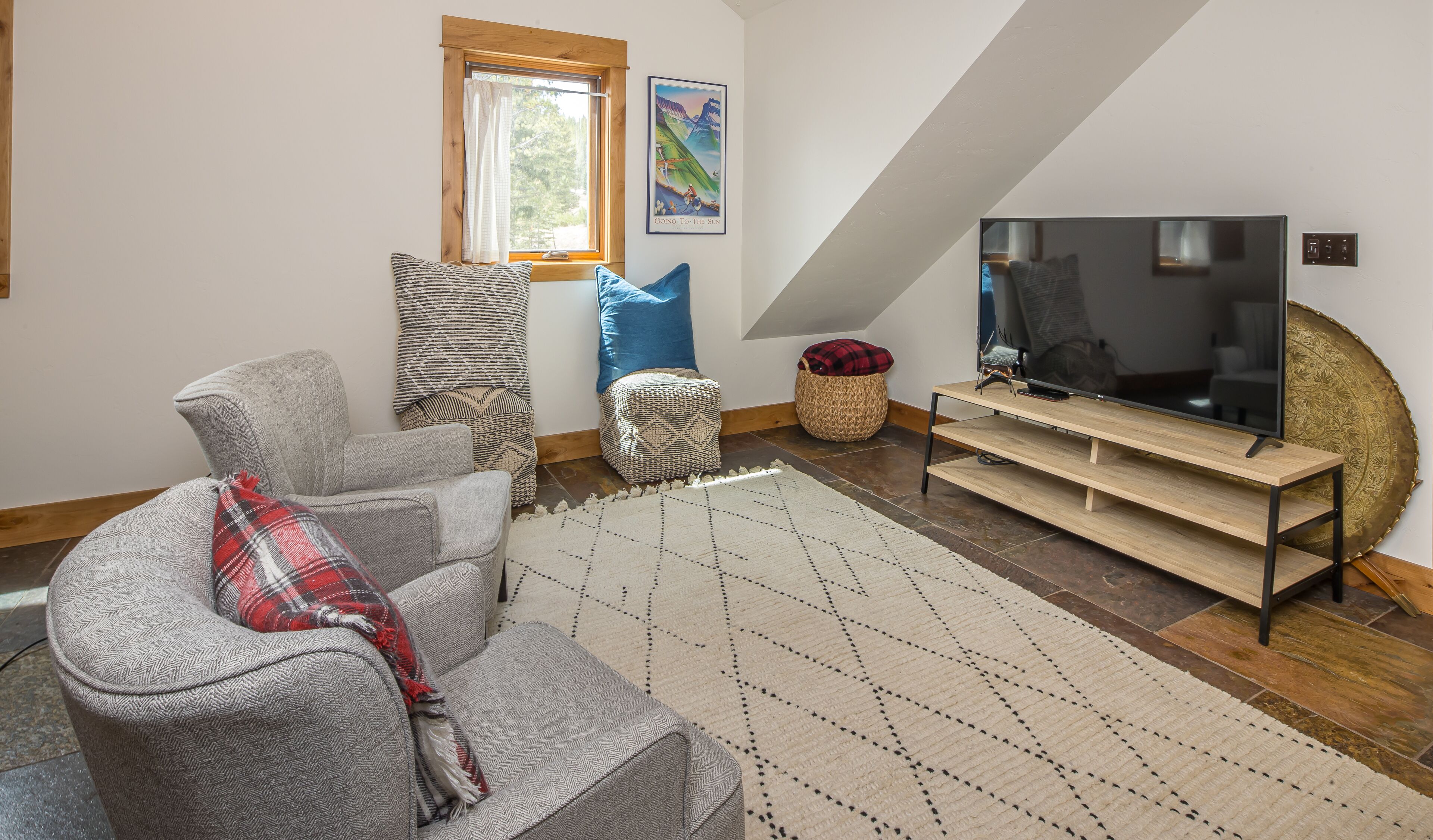 A living room with a grey sofa, patterned cushions, a large flat-screen TV on a wooden stand, a woven basket, and a window with curtains.