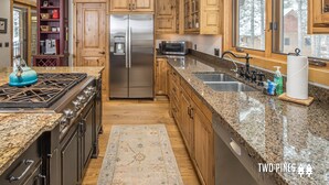 A kitchen with wooden cabinets, a stainless steel refrigerator, a gas stove, granite countertops, and a window with a view of trees. - Gorgeous Home! Private Deck, Hot Tub, Stunning Views, 15 Min Drive to  Resort! (Big Sky)