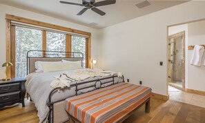Quarto com cama, mesa de cabeceira, luminária e vista para árvores através de grandes janelas.