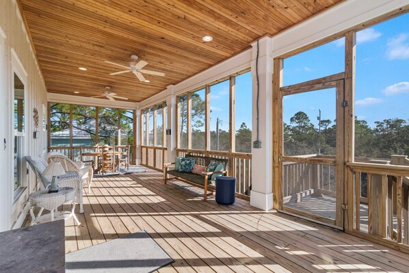 Relax in a serene outdoor space with a wooden deck, wicker chairs, and a bench, surrounded by large windows and doors offering breathtaking views.