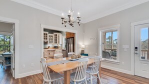 Une salle à manger spacieuse, avec une table en bois et des chaises blanches, un lustre, et une vue sur le jardin à travers de grandes fenêtres.