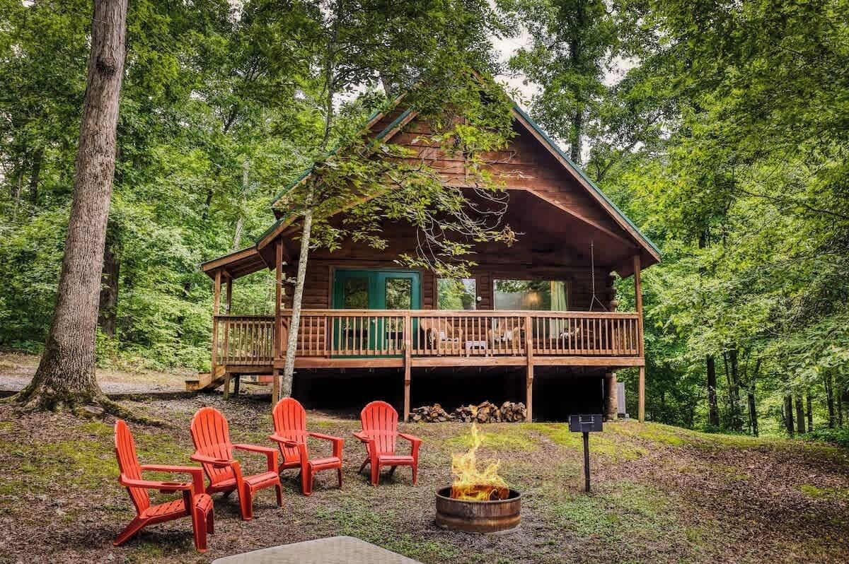 A cabin with a deck, surrounded by trees, and a fire pit with chairs.