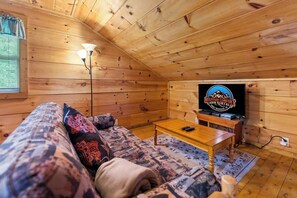 A cozy cabin interior with a wooden ceiling, a sofa with patterned cushions, a television on a stand, and a lamp.