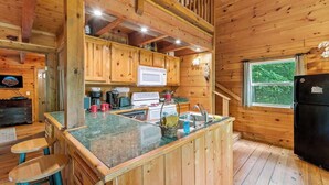 L’intérieur d’un chalet en bois comprend une cuisine dotée d’un comptoir en granit, de meubles en bois, d’un réfrigérateur noir et d’une fenêtre donnant sur un paysage verdoyant.