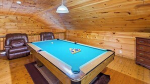Une table de billard avec une bille blanche et un jeu de billes, deux fauteuils en cuir, un plafond en bois et un luminaire suspendu.