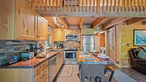Una cocina moderna con armarios de madera, una isla central, electrodomésticos de acero inoxidable y una zona de estar con sofá y mesa.