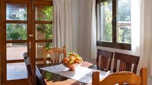 Uma mesa de jantar com uma tigela de frutas, cadeiras de madeira e uma janela com cortinas.