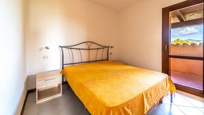A single bed with a metal frame and a yellow bedspread in a room with white walls and a window. - Appartamenti Le Carrube vicini al mare (San Teodoro)
