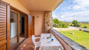 A balcony with a table set for two, a stone wall, and a view of a green landscape and road. - Appartamenti Le Carrube vicini al mare (San Teodoro)