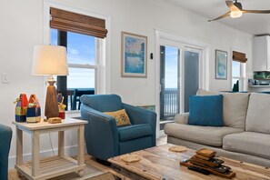 A living room with a blue armchair, a beige sofa, a wooden coffee table, and a lamp. There are windows with blinds, a ceiling fan, and artwork on the walls.