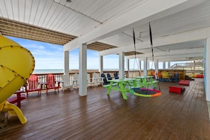 Une terrasse extérieure couverte avec un plancher en bois, des fauteuils Adirondack aux couleurs vives, une table de pique-nique verte et un toboggan jaune.