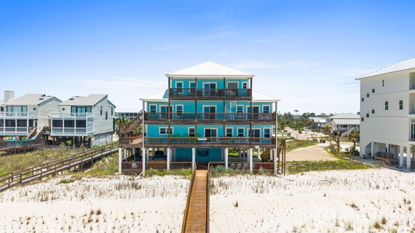 A multi-story beachfront building with balconies, a wooden boardwalk leading to it, and a sandy beach in the foreground.