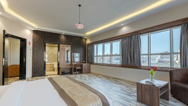 A hotel room with a large bed, a desk, a chair, a sofa, and a view of the city through large windows. - Luma Serviced Apartments (Madinah)