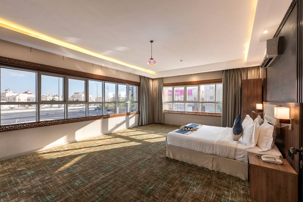 A hotel room with a large bed, a bedside table with a lamp, a view of the city through large windows, and a patterned carpet.