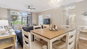 A spacious living room with a wooden dining table, comfortable seating, a ceiling fan, and a large window offering a view of the outdoors.