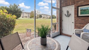 A patio with a view of a grassy field and buildings, a potted plant, and wicker furniture.