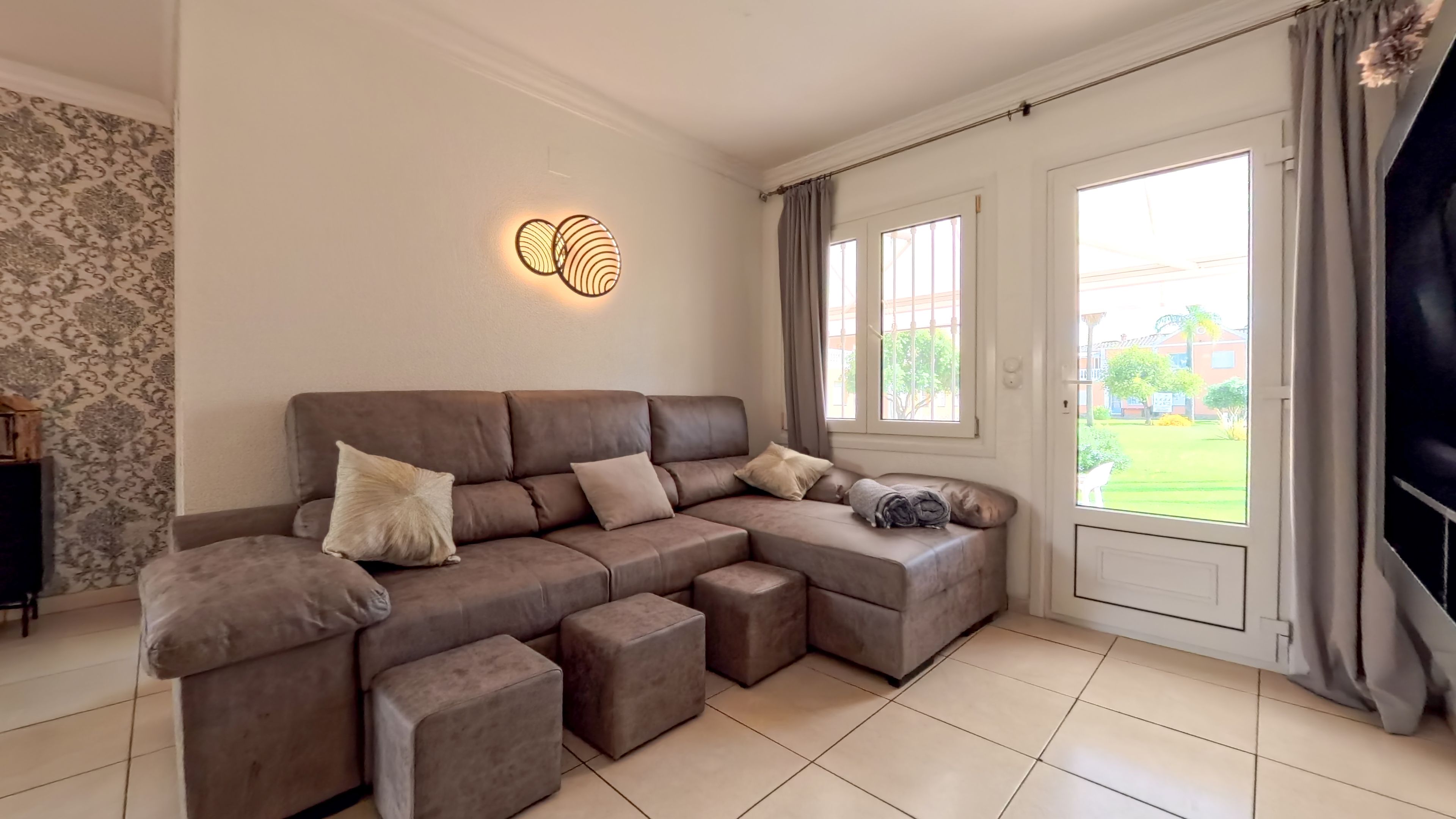 A modern living room with a sectional sofa, ottomans, a decorative wall light, and a view of a courtyard through a glass door.
