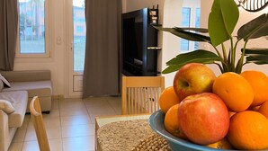 A living room with a sofa, a TV, a dining area with a table and chairs, and a bowl of fruit on the table. - Solea Mare by Midelisa (oliva nova golf)