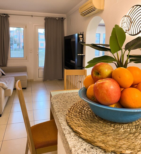 A living room with a sofa, a TV, a dining area with a table and chairs, and a bowl of fruit on the table. - Solea Mare by Midelisa (oliva nova golf)