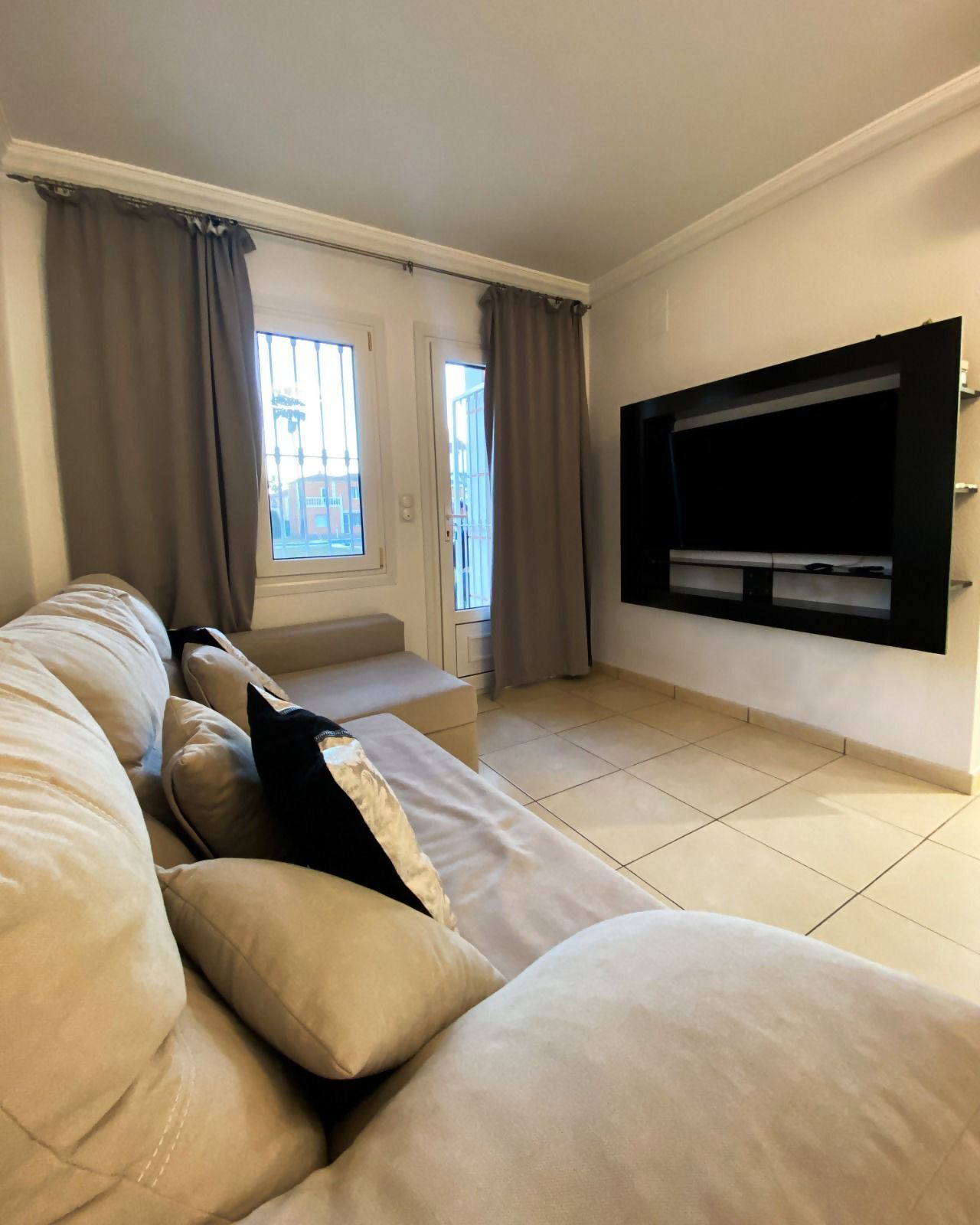 Una sala de estar con un sofá color beige, un televisor de pantalla plana montado en la pared y un ventanal con cortinas.