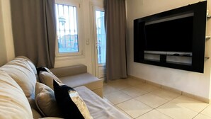A living room with a beige sofa, a flat-screen TV mounted on the wall, and a window with curtains.