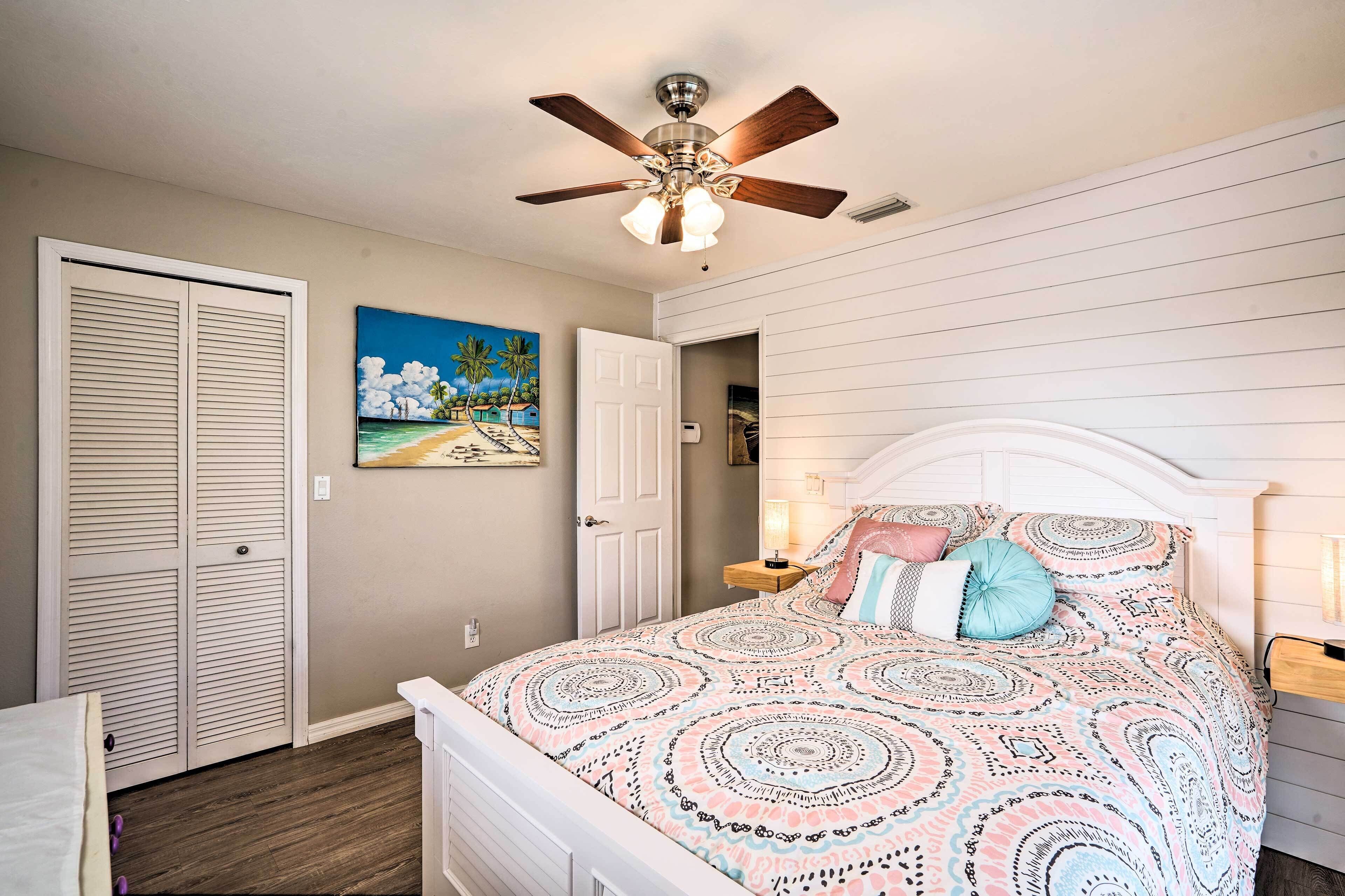 A bedroom with a bed, a ceiling fan, a painting, and a door.