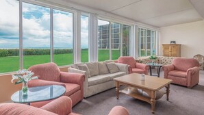 A spacious living room with large windows, a beige sofa, two pink armchairs, a wooden coffee table, and a small glass table with a vase. - Large Beach Condo!! Amazing Ocean Views!! Hutchinson Island, Jensen Beach, FL (Jensen Beach)
