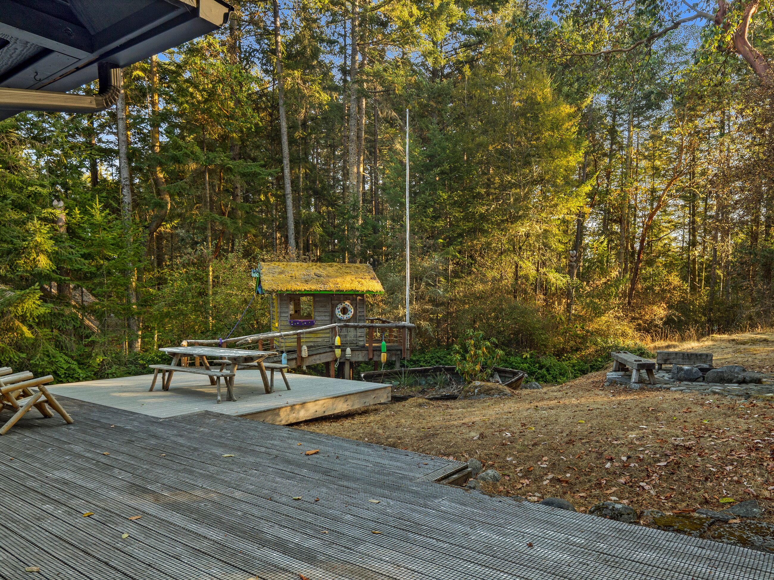 Una terraza de madera con mesas de picnic, una pequeña construcción de madera con techo de paja y un fondo de bosque.