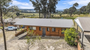 A single-story brick building with a metal roof, a covered carport, and a small garden area with a tree and shrubs. - Cuttagee Beach Cottage 6 - Tathra Bermagui Rd 3521 (Barragga Bay)