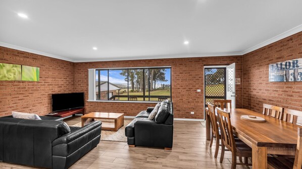 A spacious living room with a brick wall, a flat-screen TV, a black leather sofa, a wooden dining table with chairs, and a large window offering a view of trees and a house.
