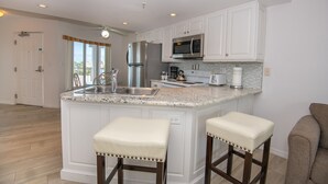 A kitchen with white cabinets, a stainless steel refrigerator, a microwave, and a granite countertop. There are two bar stools with white cushions. - Luxury Beach Escape | Steps to Sand (St. Pete Beach)