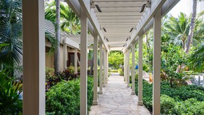 A covered walkway with stone flooring, surrounded by lush greenery and palm trees. - The Watermark (WM1102) Luxury 11th-floor 2 BDR, balcony, AC, remote work (Honolulu)