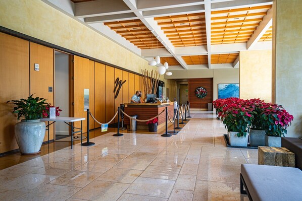 A hotel lobby with a reception desk, wooden paneling, potted plants, and a tiled floor.