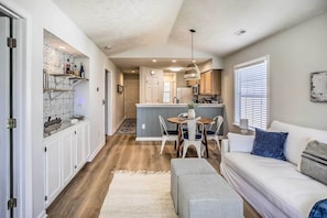 A modern kitchen with white cabinets, a dining area with a table and chairs, and a living room with a sofa and a bench.