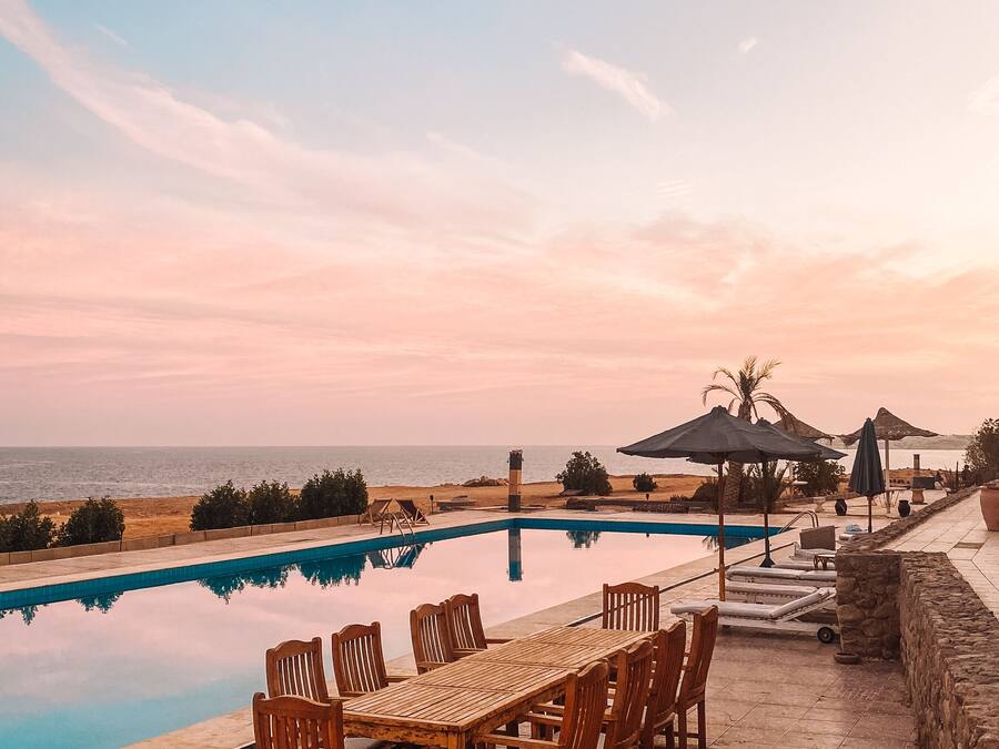 Un espace aménagé au bord de la piscine, avec une table et des chaises en bois, un mur en pierre et une vue sur la mer au coucher du soleil.