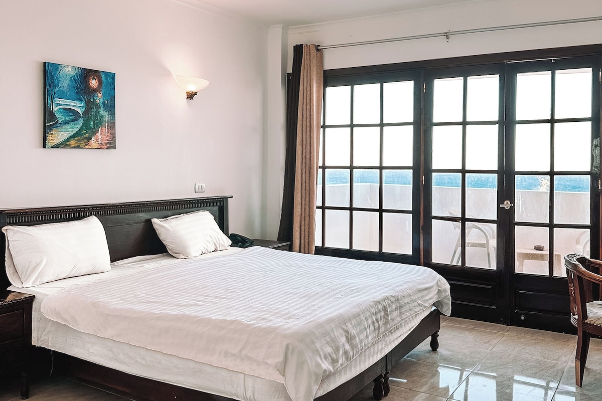A hotel room with a large bed, a nightstand, a painting, a window with curtains, and a view of the outside.