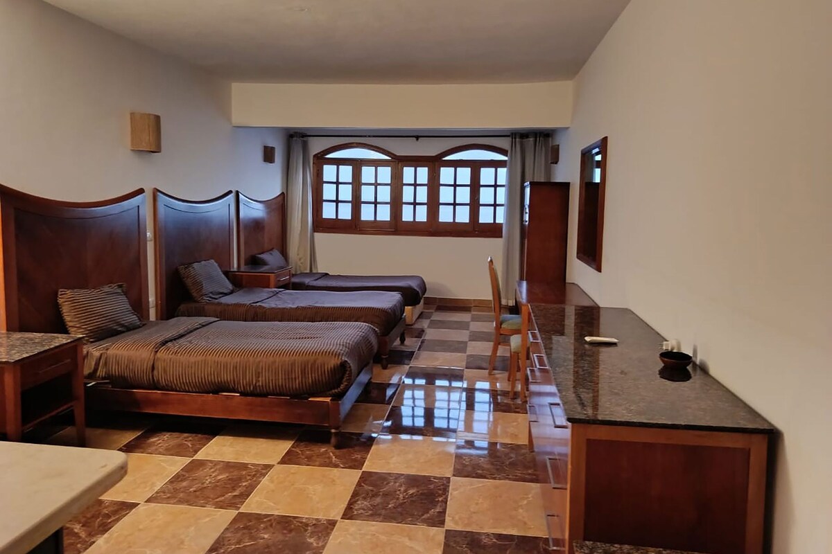 A hotel room with a checkered floor, two wooden beds, a desk, and a window with a view.