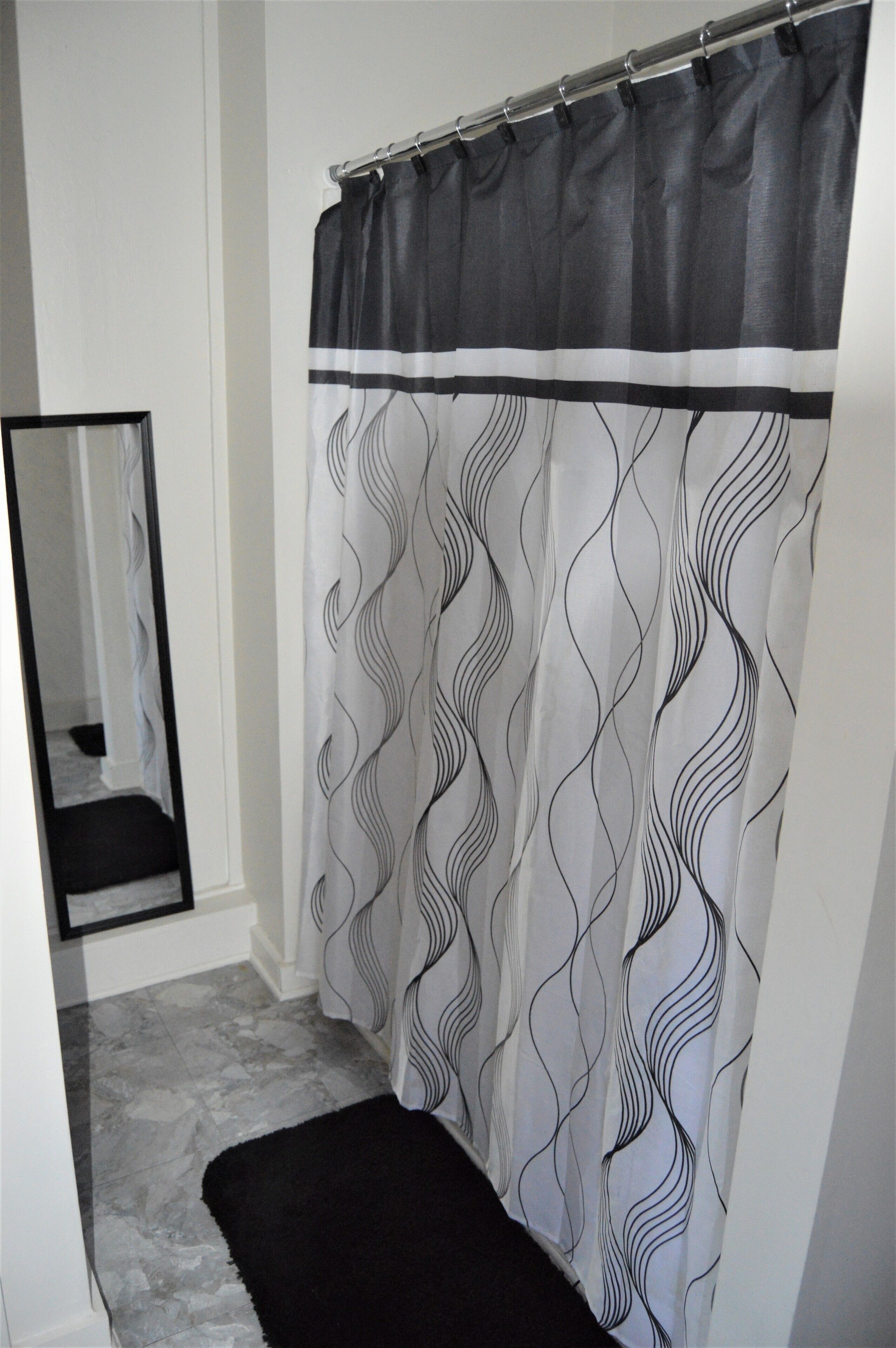 Une salle de bain avec un rideau de douche orné d’un motif ondulé, un tapis noir et un miroir au mur.