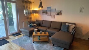 A living room with a sectional sofa, a coffee table, and a lamp. There are framed pictures on the wall and a window with a view of the outdoors.