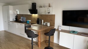 A modern kitchen with white cabinets, a wooden countertop, and a black range hood. There are two black bar stools and a large flat-screen TV mounted on the wall.