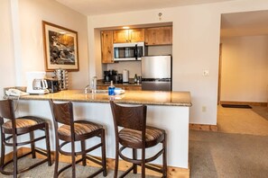 Una cocina con microondas, refrigerador y gabinetes de madera. Hay tres taburetes de bar y una encimera con un lavamanos.