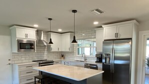 Una cocina moderna con armarios blancos, electrodomésticos de acero inoxidable, una isla central con taburetes e iluminación colgante.