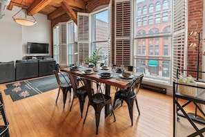 A dining area with a wooden table, black chairs, and a view of a brick building through large windows. - Luxe Loft Downtown (Indianapolis)