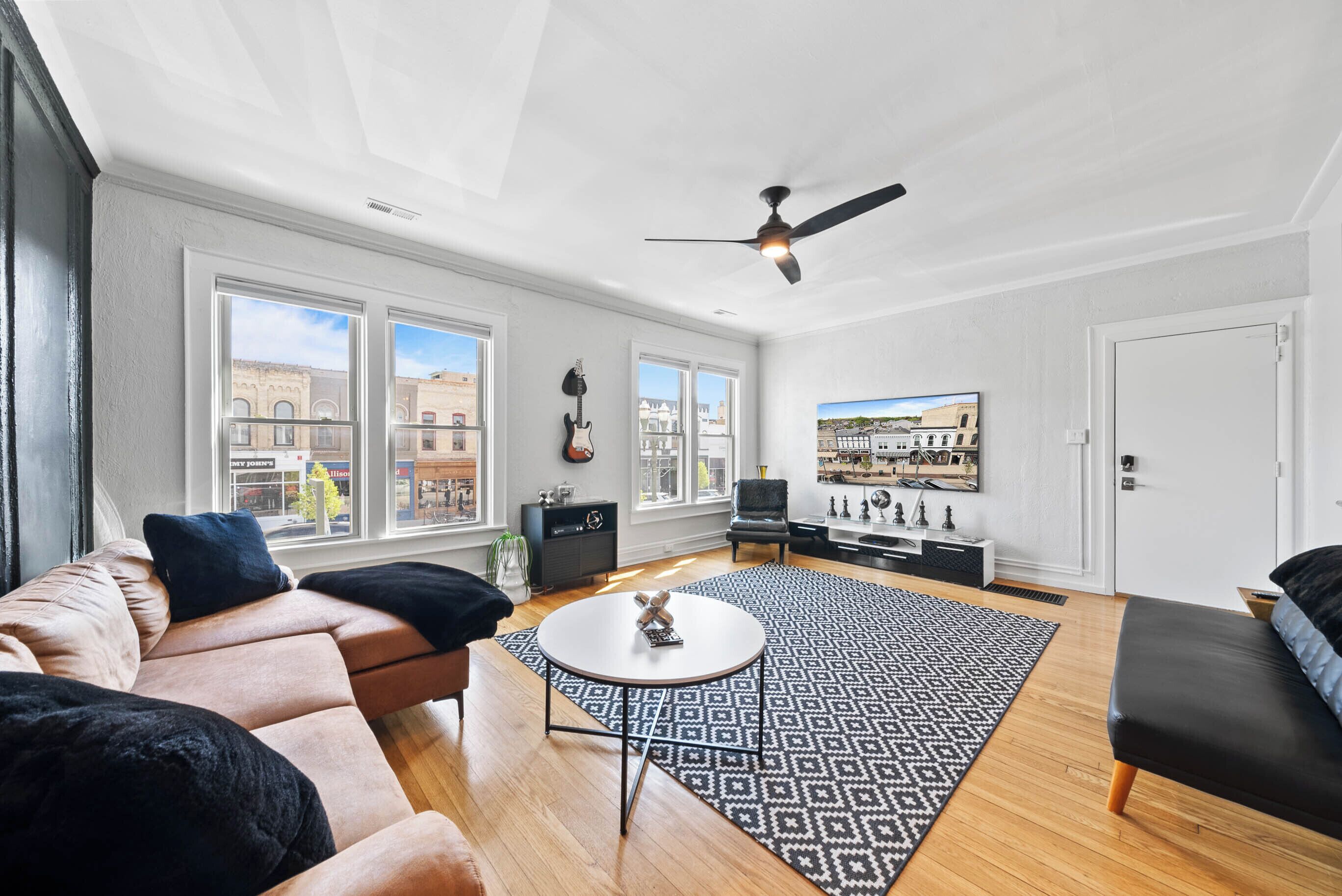 A spacious living room with a large window, a ceiling fan, a sofa, a coffee table, and a patterned rug.