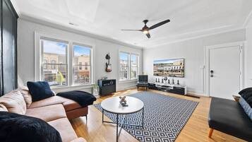 A spacious living room with a large window, a ceiling fan, a sofa, a coffee table, and a patterned rug.